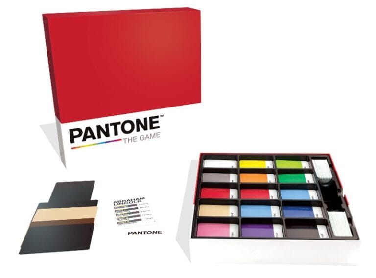 The box for "Pantone: The Game" with a red lid takes center stage, perfectly showcasing this vibrant card game. Displayed alongside the open box, colorful cards and components hint at endless creativity. Get ready—it's releasing tomorrow!