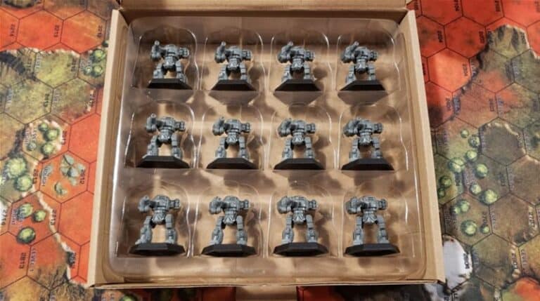 An open box set contains twelve gray miniature UrbanMech robots, aligned in a 3x4 grid. The background features a hexagonal-patterned game board with earthy tones, reminiscent of a Catalyst Game Labs creation.