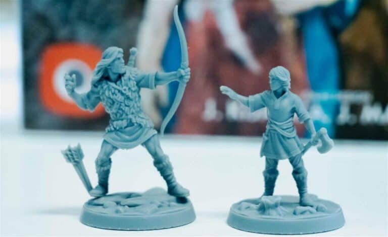 Two gray miniatures of medieval warriors stand on a tabletop, reminiscent of Thorgal adventures. One wields a bow and is dressed in fur, while the other holds an axe. A blurred colorful background features indistinct text and imagery, evoking scenes from a captivating board game.