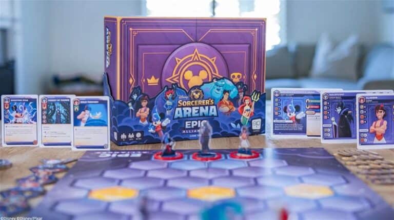 A game setup for the Disney Sorcerer's Arena: Epic Alliances features character cards and miniatures. The game box with colorful artwork is in the background, surrounded by hexagonal board pieces and various cards on a wooden table.