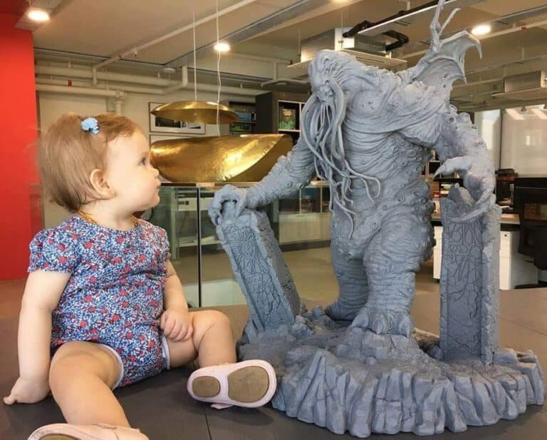 A toddler in a floral outfit sits on a table, gazing curiously at a large, detailed statue of Cthulhu from CMON's Death May Die. The setting is an indoor space with modern decor, creating an intriguing contrast between innocence and myth.