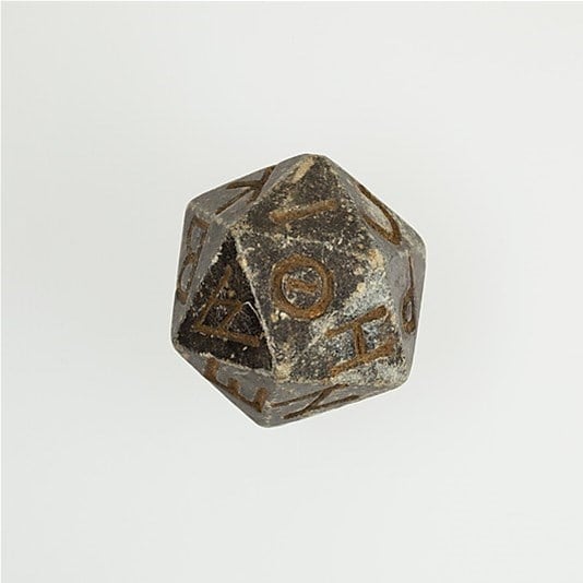 An ancient, worn icosahedron die, possibly thousands of years old, with faded symbols on each face, set against a plain white background. This unique dice appears to be made of a dark, stone-like material.