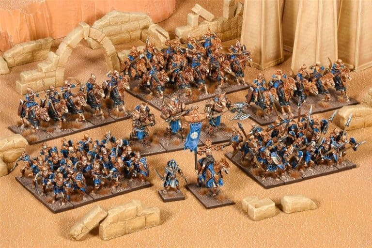 A detailed tabletop wargame diorama from Mantic Games showcases an army of miniature fantasy warriors from the Empire of Dust. The figures, in blue and gold armor, wield weapons across a desert-themed landscape with ruins and sand-colored terrain.