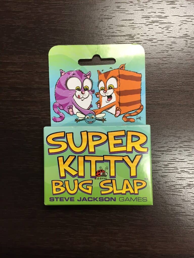 A card game package for "Super Kitty Bug Slap" by Steve Jackson Games, noted in a TGN Review, showcases artwork with a purple and an orange cat eagerly reaching for a small bug. The packaging rests elegantly on a dark wooden surface.