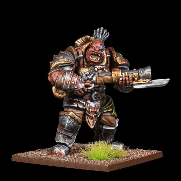 A painted figurine of a fantasy warrior orc from the Kings of War series, wielding a large gun with an attached blade. The orc, clad in armor and sporting a mohawk, stands on a textured base with patches of grass. The background is completely black.