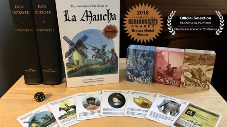 Box of the "La Mancha" card game sits proudly on a table, its contents spread open with cards and a die. A Kickstarter success, it’s accompanied by two Don Quixote books. The image proudly notes its bronze medal from the 2018 International Serious Play Awards and official selection.