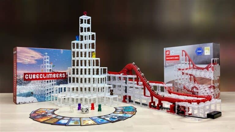 A tabletop game set featuring a tall tower of clear blocks, red tracks with small cars, and a fan-like array of colorful cards. Two boxes labeled "Cube Climbers," showcasing its vibrant style, and "Nanoblocks" are in the background, making this board game an exciting adventure for all ages.