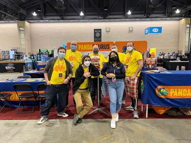 A group of six people, some sporting matching yellow Pandasaurus-themed shirts, pose together at a bustling convention booth. The banner reads "Panda," with various items enticingly displayed on tables. In the background, masked attendees wander through the lively scene.