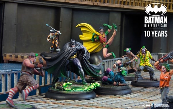 Miniature figures from the Batman universe, including Batman and Robin, are displayed on a platform surrounded by other characters. The scene captures an action-packed moment from the Knight Models' Batman Miniature Game, celebrating its 10th anniversary with exciting new releases.