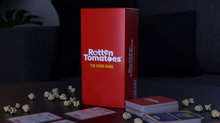 A red box labeled "Rotten Tomatoes: The Card Game" by Cryptozoic sits on a table amid scattered popcorn kernels and an array of cards. Some cards are spread out, with a cozy dark sofa and a lush plant providing the perfect backdrop.
