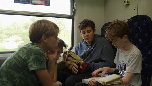 Four boys sit on a train, engaged in reading and conversation. The one on the far right is focused on a book about D&D, while the others listen and look out the window. Their session zero chat feels like brainstorming for a Kickstarter campaign—calm yet full of excitement and potential.