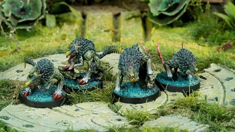 Miniature models of wolf-like creatures, akin to the Hungries from Corvus Belli's Infinity universe, feature dark scales and red accents. Posed on rocky bases with a water effect, they are set on a grassy terrain lush with vegetation and scattered rocks.