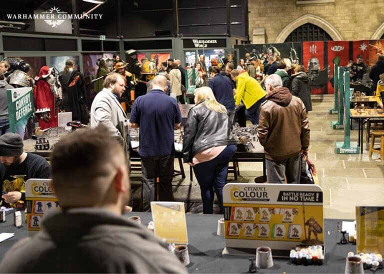 A bustling tabletop gaming convention fills the brightly lit hall, where attendees eagerly engage at various booths. It's Open Day 2023 at Warhammer World, with enthusiasts browsing exclusive Warhammer merchandise and admiring the intricately decorated tables on display.