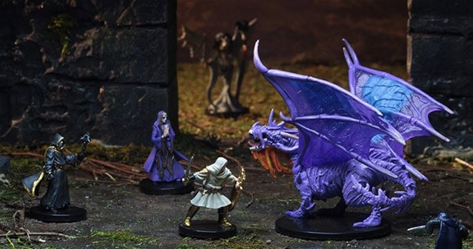 In the dark, dungeon-like environment of the Ruins of Lastwall, a group of miniature fantasy figures, including two robed characters and an armed adventurer from Pathfinder Battles, confronts a menacing purple dragon with its wings spread wide.
