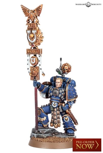 A detailed miniature of a blue-armored Ultramarines Space Marine serving as a Standard Bearer. The figure boasts intricate details and a gold decoration atop the staff, crafted by Forge World. The rocky base supports a "Pre-order Now" sign in the bottom right corner.