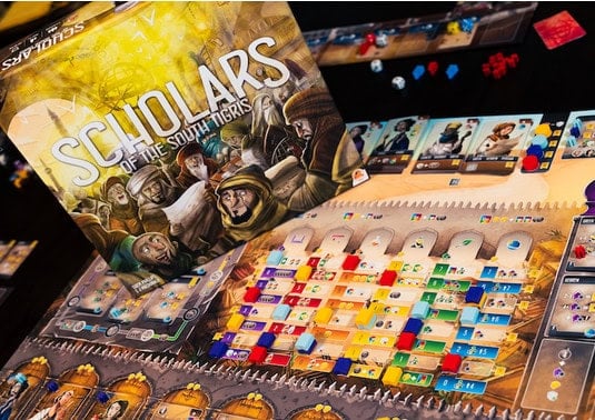 A board game titled "Scholars of the South Tigris" is set up. The colorful board, integral to this strategic dice bag-building game, features various tracks and components, with pieces in use. The box art depicts medieval scholars against an amber-lit background, hinting at its Kickstarter roots.