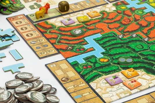 Close-up of a colorful board game featuring terrain reminiscent of the Alpujarras, with red and green areas and a blue river. Several game tokens, tiles, and stacks of coins are scattered around the board. A yellow pawn from the Kickstarter Campaign edition and a die are visible on the board.