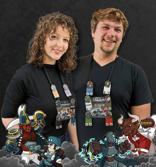 A smiling man and woman in black shirts with lanyards, each holding several cards from the Wearable Trading Card Game. Surrounding them are illustrated fantasy characters like a warrior, a soldier, and a minotaur against a dark background. It's an adventure of Worn Wanderers you can Play Anywhere.