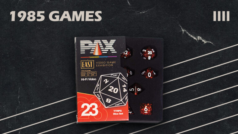 A set of special edition dice is displayed in an open box with the label "PAX East Video Game Exhibition." Featuring retro-themed graphics and the number "23," this Penny Arcade collaboration sits against a backdrop of diagonal white lines with the text "1985 GAMES.