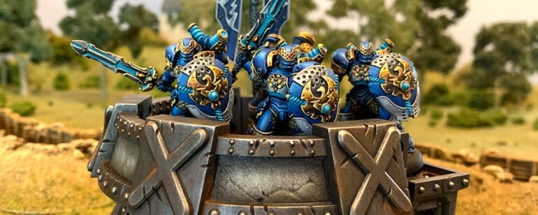 Four painted miniature warriors in ornate blue and gold armor stand atop a fortified stone structure, resembling intricate bunkers. They hold shields and swords, poised for battle against the lush, green landscape—a stunning piece from Privateer Press that highlights every terrain feature beautifully.