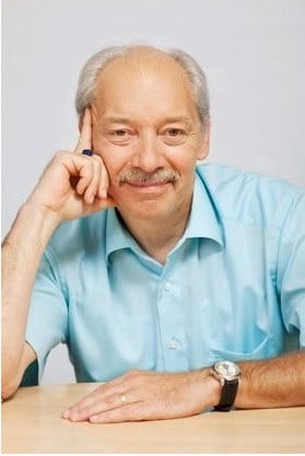 A smiling man with a mustache, wearing a light blue shirt, sits at a table resting his head on his hand. With a wristwatch and ring, he exudes the relaxed wisdom of someone who might be pondering strategies for Settlers of Catan. The background is plain and light-colored.