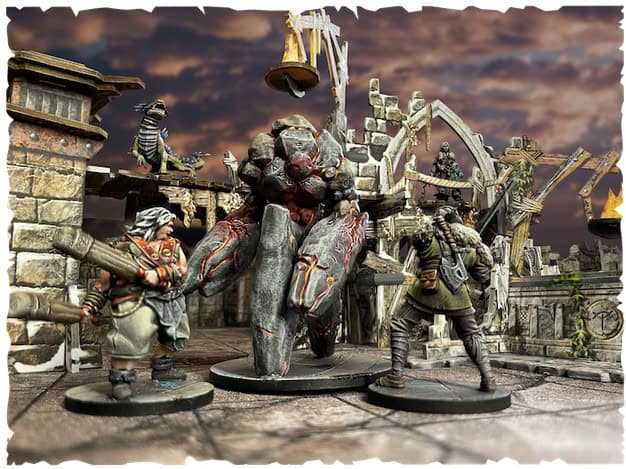 In a dramatic tabletop scene, two miniature figures confront a large armored creature amid the ruins of "Dungeons of Enveron." The cloudy sky adds tension, framed by ancient architecture and a flaming brazier. Discover more in the Maladum Kickstarter Campaign.