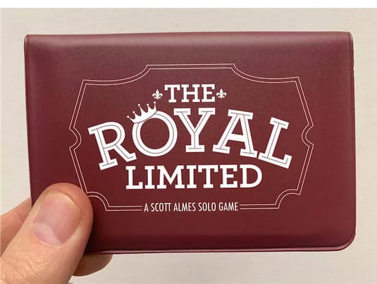 A hand holds a maroon rectangular box labeled "The Royal Limited," a Scott Almes solo wallet game that evokes the elegance of luxury train travel. The design features a crown emblem above the text, making this Kickstarter gem truly captivating.
