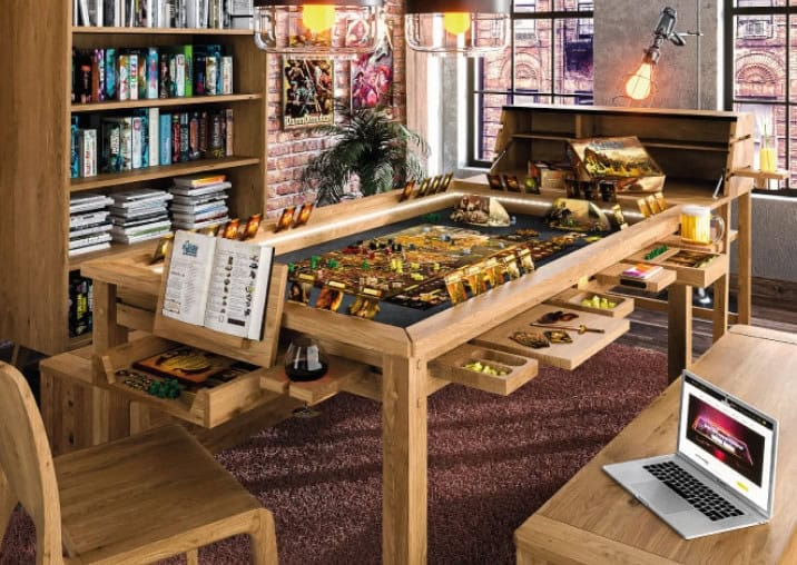 A cozy gaming room features a central Geeknson table set up for a board game. Shelves filled with books and games line the wall. A laptop is open on a nearby bench, glowing under warm lighting that creates an inviting Kickstarter ambiance.