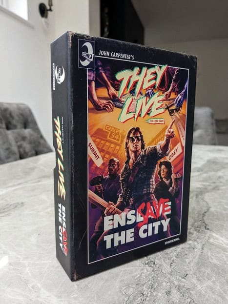 A Kickstarter board game box titled "They Live" sits on a table. The cover features an illustration of people, some wearing sunglasses, surrounded by slogans like "Submit" and "Obey." The tagline "Enslave the City" is prominently displayed.