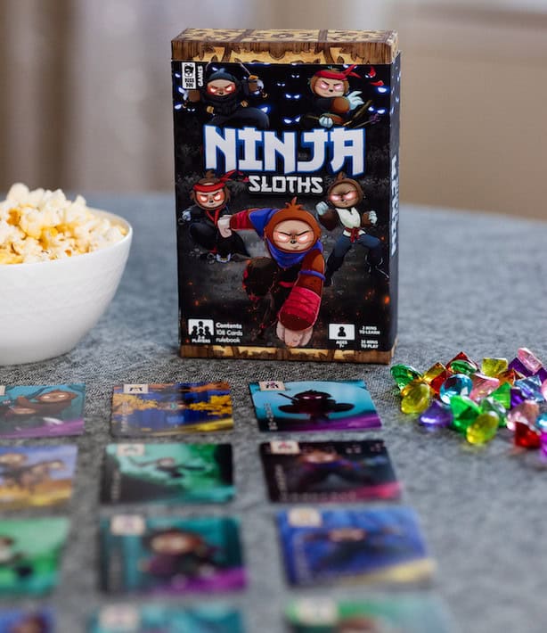 A tabletop setup features the "Ninja Sloths" card game, fresh from its successful Kickstarter campaign. The game box displays cartoon sloth ninjas, with a bowl of popcorn and colorful tokens scattered among the character cards on a textured surface.