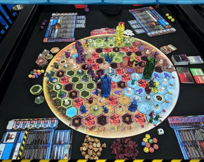 A complex board game setup on a table, featuring a hexagonal-tiled map from Star Reach Games, colorful tokens, dice, cards, and several pieces scattered around. Various thematic elements suggest a strategy or adventure game immersed in the Cysmic universe.