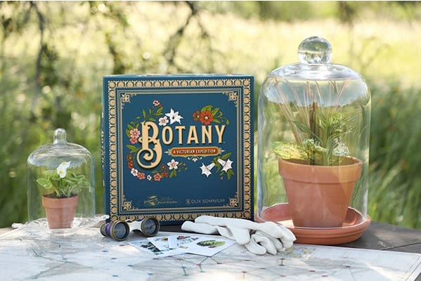 A book titled "Botany: A Victorian Expedition" lies on a table alongside two glass cloches covering potted plants, a pair of gloves, a magnifying glass, and plant illustrations. This captivating display invites you into the fascinating world of Victorian plant hunters against a grassy outdoor backdrop.