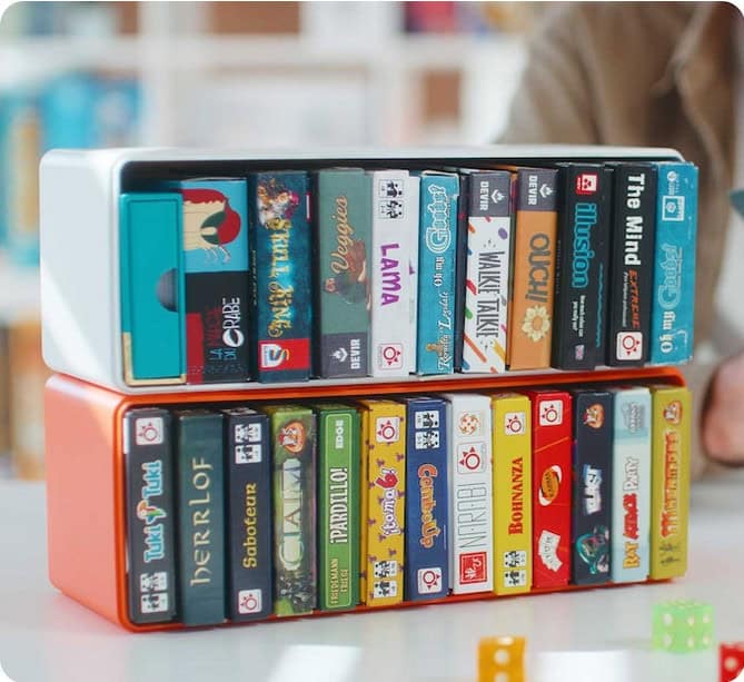 A collection of small, compact board and card game boxes neatly organized on two shelves showcases an ingenious storage solution. Various game titles and colorful designs are visible, hinting at successful Kickstarter campaigns. A pair of dice can be seen in the foreground.