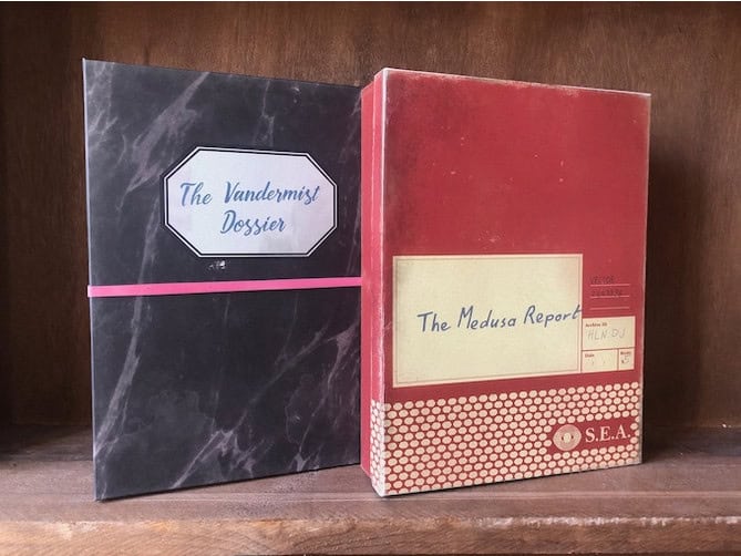 Two files stand on a wooden shelf. One is labeled "The Vandemist Dossier" with a black and white cover. The other, "The Medusa Report," sits in a red binder with a cream label marked as "SEA." This intriguing pair hints at Cold War secrets worthy of their own Kickstarter campaign.