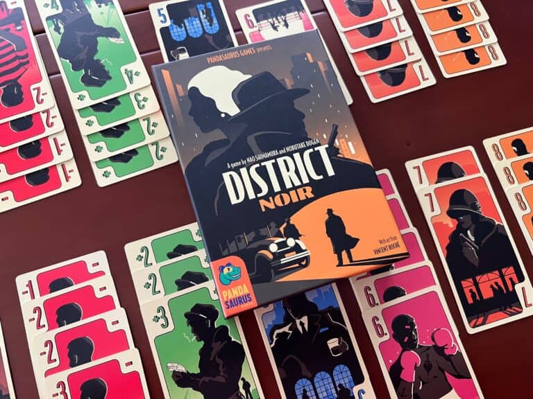 A card game titled "District Noir" awaits eager players on a table surrounded by colorful cards. The box, available for preorders, features a detective's silhouette against a cityscape in dark and orange tones. Cards showcase various images in red, green, and other strategic battle colors.