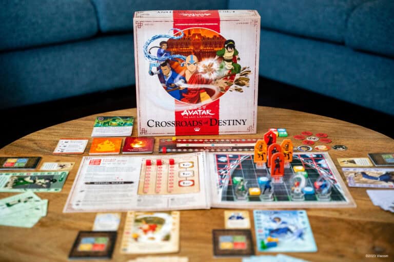 A board game titled "Avatar: Crossroads of Destiny" by Funko Games is displayed on a table. The setup includes a game board, cards, and various pieces with illustrations from Avatar: The Last Airbender. The game's box features animated characters in dynamic poses, highlighting its co-op strategy elements.