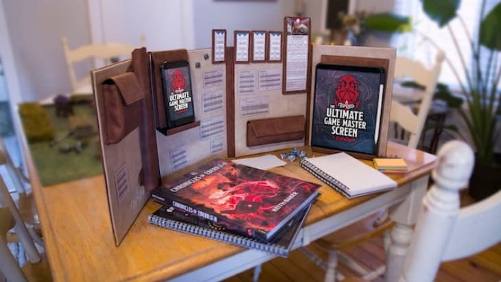 A wooden tabletop is set with the Kickstarter-funded Ultimate Game Master Screen, surrounded by notebooks and role-playing game books. Dice and a pencil lie nearby, ready for adventure. Chairs are in the background, and plants add a touch of nature to the room.