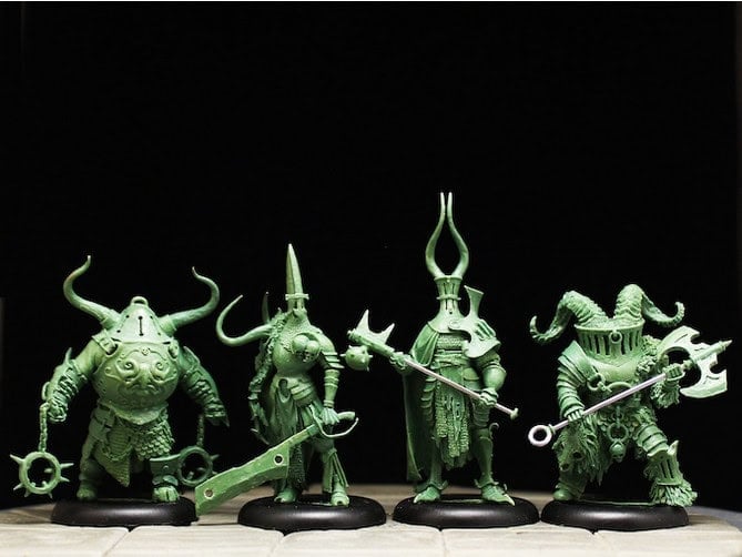 Four green, intricately detailed miniatures from Westfalia Publishing stand on a table. Each figure is wearing armor with elaborate designs, horns, and helmets, holding weapons like axes and staffs. The background is a dark black, highlighting the vibrant green figures.