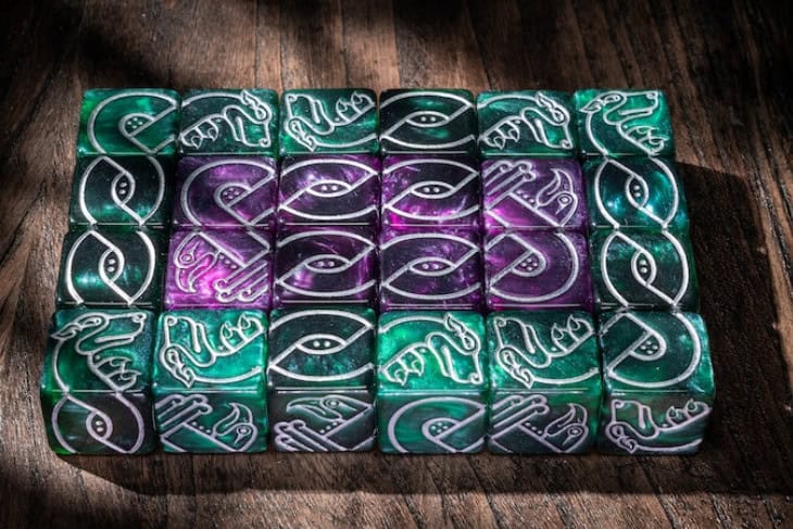 A set of intricately designed Celtic Animal Knot Dice is arranged in a grid on a wooden surface. The dice feature green and purple hues with silver, swirling patterns resembling an eye motif. Sunlight casts diagonal shadows across the scene, sparking creativity for any Kickstarter project.