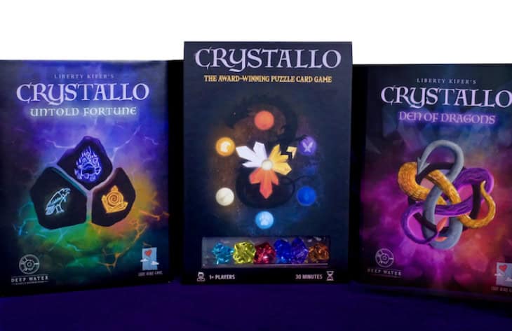 Three Crystallo board game boxes are displayed: "Untold Fortune" adorned with crystal illustrations, "Crystallo" showcasing hexagonal colored gems, and "Den of Dragons" featuring interlocking golden and purple rings. The dark, gradient background adds an aura of mystery.