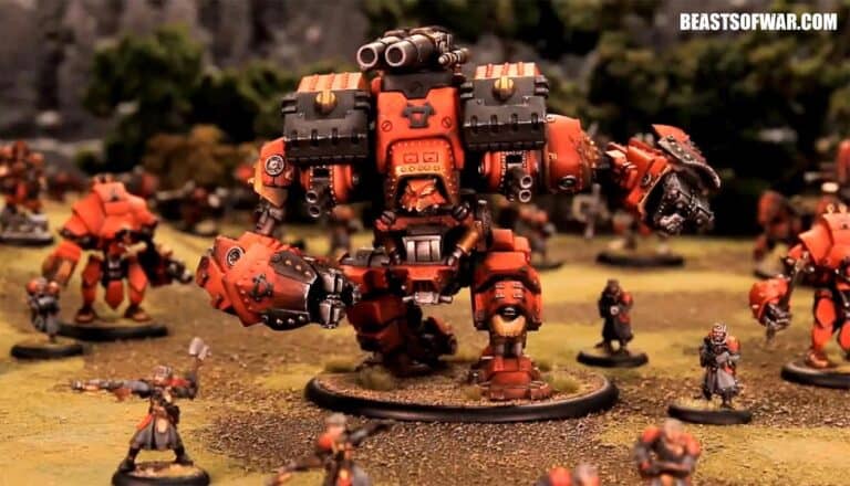 A large, red mech figurine by Privateer Press stands prominently on a wargaming table, surrounded by smaller figures. The scene is set outdoors, with grassy terrain and miniature trees in the background, capturing the essence of legacy model production.