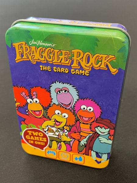 A vibrant tin box for "Fraggle Rock: The Card Game" by River Horse showcases cartoon characters with colorful hairstyles, all smiling joyfully. The text proudly announces "Two Games in One!" set against a playful purple background.