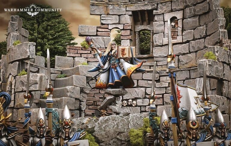 In this fantasy miniature scene, an elf-like figure in ornate blue and white armor stands atop a ruined stone fortress, reminiscent of Warhammer: The Old World. Armored soldiers with blue banners surround them, set against a backdrop of trees and a cloudy sky.