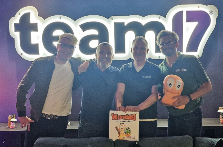 Four people stand together in front of a brightly lit "team17" sign. Two proudly hold the "Worms The Board Game" box, likely a collaboration with Mantic Games, while another holds a plush toy resembling a character from the game. They are all smiles, clearly celebrating something special.