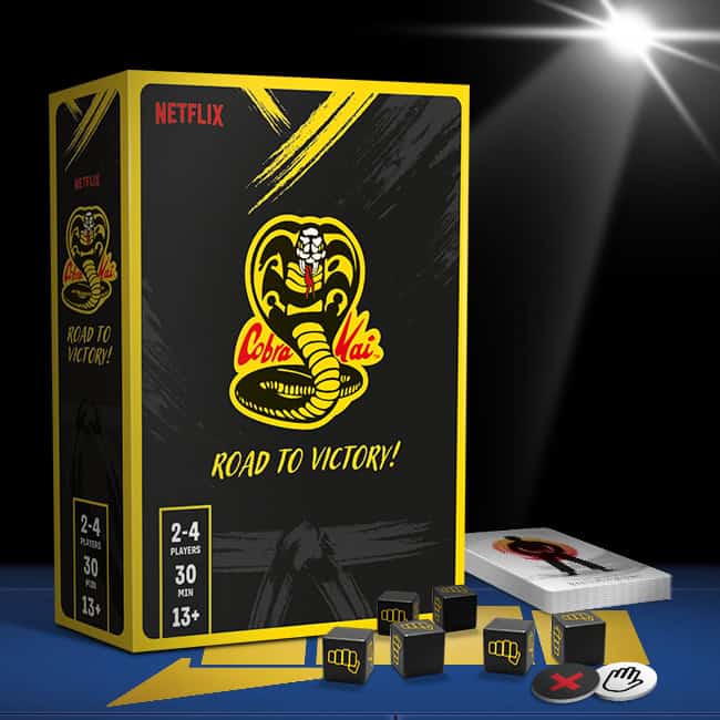 The box cover and components of "Cobra Kai: Road to Victory," a thrilling board game published by Asmodee, feature a striking yellow cobra logo, dice, playing cards, and tokens. Suitable for 2-4 players aged 13 and up, it promises an exciting 30-minute playtime.