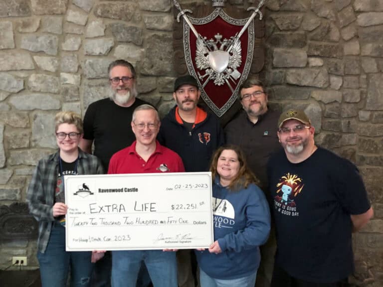 A group of seven people stands indoors before a stone wall at Ravenwood Castle, holding a large check for $22,251.51 made out to "Extra Life," dated 02-25-2023. Behind them, a decorative shield and the atmosphere of a Charitable Gaming Event add to the excitement.