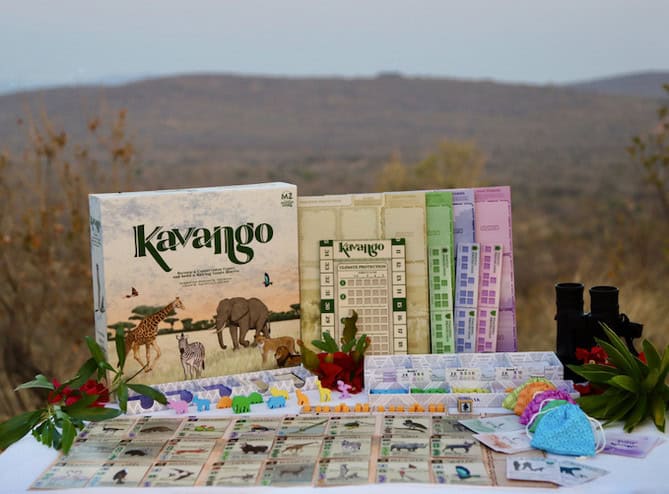 A board game titled "Kavango" is set up outdoors on a table. The game box, designed by a conservation expert, features illustrations of wildlife like elephants and giraffes. Game components, cards, and pieces are displayed, with binoculars and plants nearby against a hilly landscape backdrop.