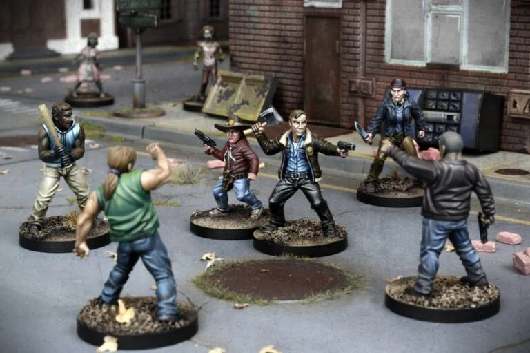 Miniature figures are arranged as if in a standoff on a street setting, evoking scenes from Mantic Games' The Walking Dead board game. They include characters with weapons and detailed urban elements like brick buildings and debris, creating an immersive experience.