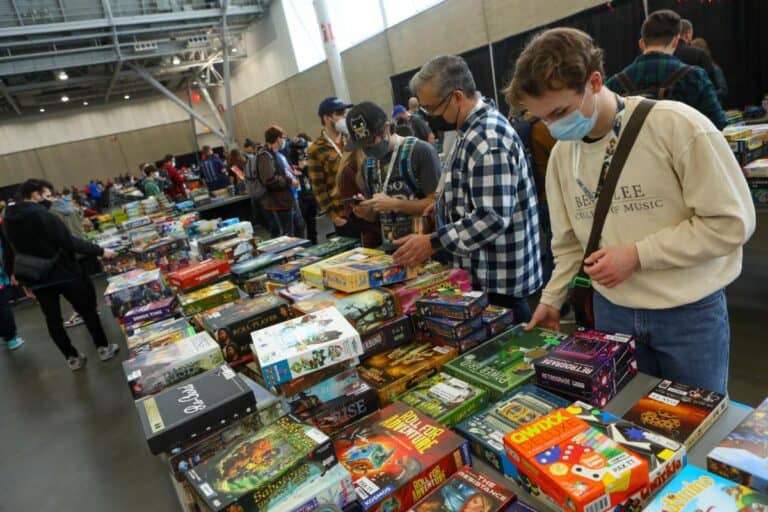 At PAX East 2024, attendees browse a large selection of board games on tables. Most are wearing casual clothes and some have masks, enjoying titles like Marvel Dice Throne. The spacious, industrial-style hall buzzes with excitement as more gamers explore the convention.