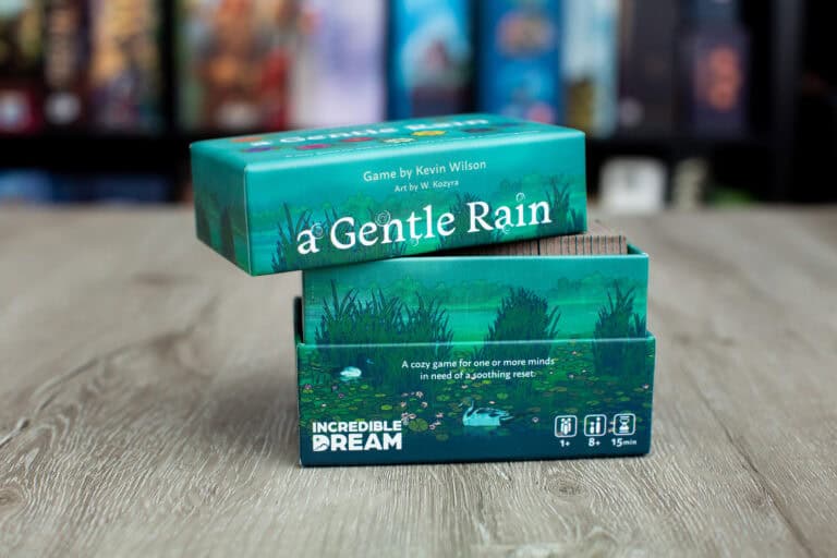 A green board game box titled "A Gentle Rain: Bloom Edition" is partially opened, unveiling artwork of a serene water scene with lily pads. Text on the box says it's for 1-2 players, ages 8+, lasting 15 mins. Created by Kevin Wilson with art by K. Kwapis, an incredible dream in gameplay awaits.